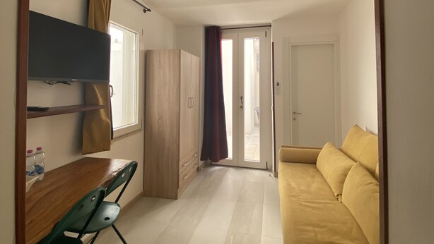 Quarto com sofá amarelo, escrivaninha de madeira com cadeira, guarda-roupa e porta de vidro deslizante que dá para uma área externa.
