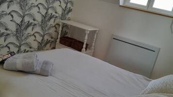 A neatly made bed with white linens and a folded towel, a small white nightstand with a basket, and a radiator on the wall.