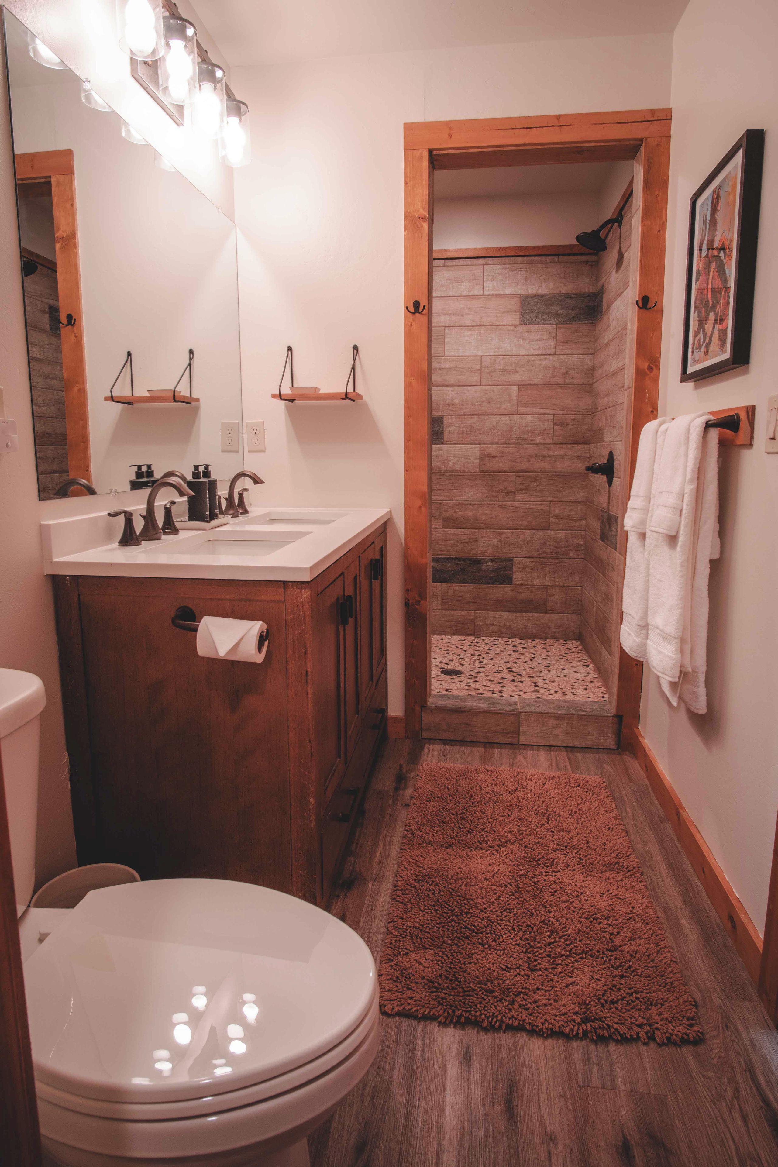 A bathroom with a wooden vanity, a white sink, a mirror with lights, a shower with a wooden door, and a brown rug.