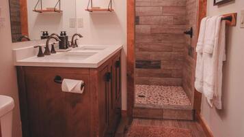 A bathroom with a wooden vanity, a white sink, a mirror with lights, a shower with a wooden door, and a brown rug.