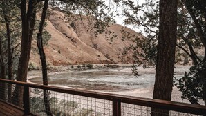 Une rivière avec une terrasse en bois et une rambarde en métal au premier plan, entourée d’arbres et d’une colline en arrière-plan.
