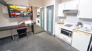 Moderni hotellihuone, jossa on taulutelevisio, työpöytä kannettavalla tietokoneella, pieni tuoli, minikeittiö, jossa on mikroaaltouuni, uuni ja pesuallas, sekä lasiovi, joka johtaa kylpyhuoneeseen.