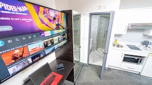 Hotellihuone, jossa on suuri televisio, jolla näkyy elokuva, kannettava tietokone, älypuhelin, suihku ja pieni keittiö.