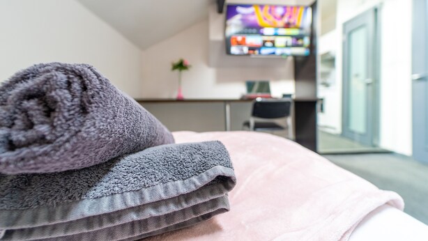 Siististi pedattu sänky, jossa on vaaleanpunainen ja harmaa peitto, pieni työpöytä tuolilla ja seinälle asennettu televisio.