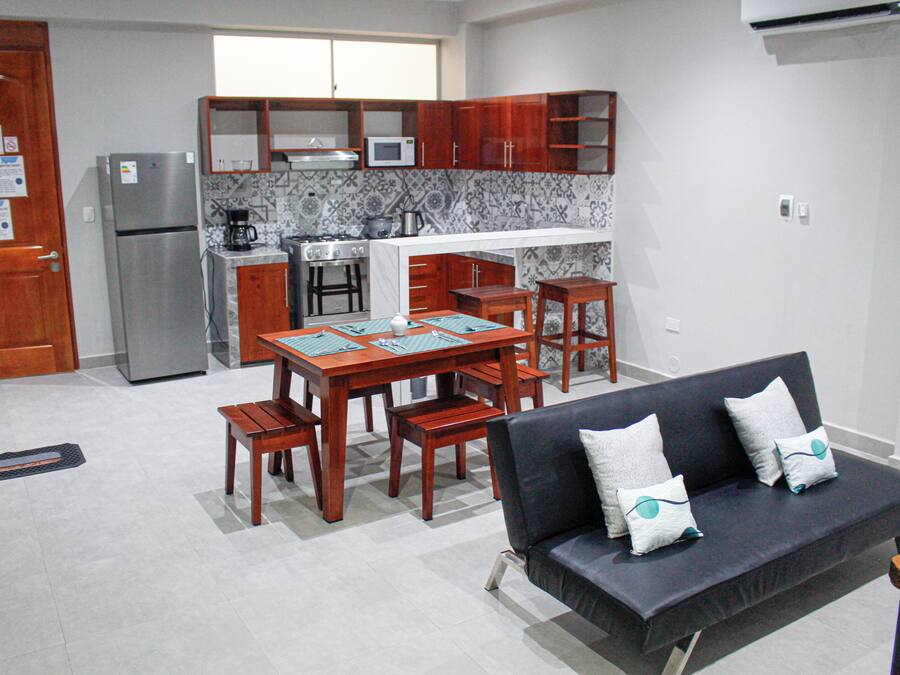 A compact kitchen with a refrigerator, stove, and microwave. There is a dining table with chairs and a sofa with cushions.
