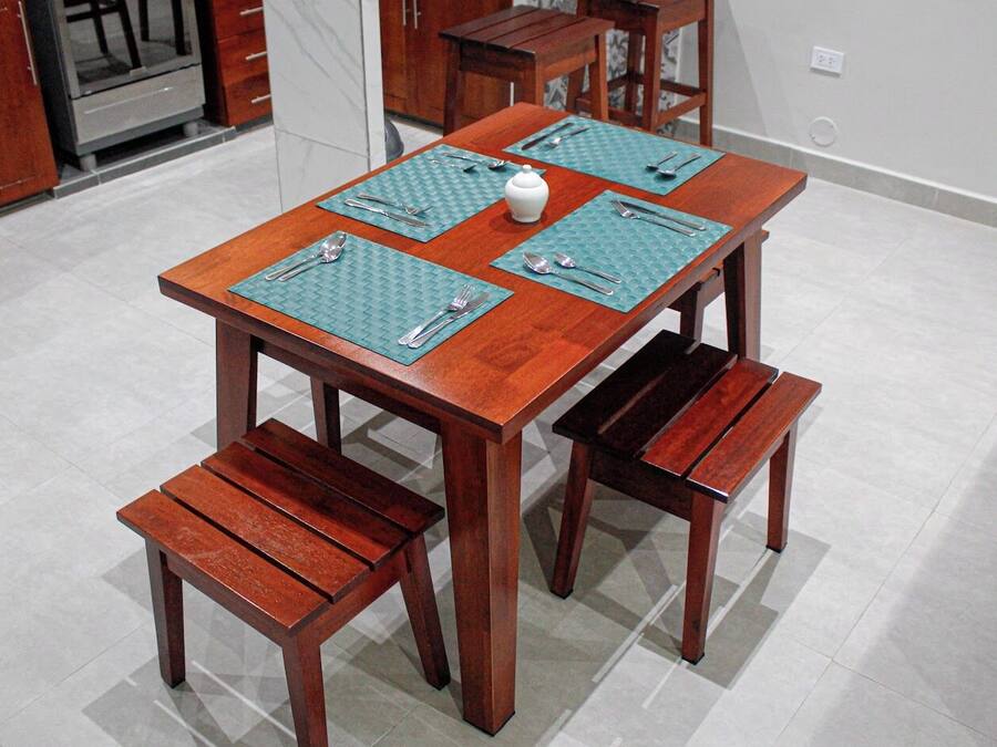 A wooden dining table with four matching benches, a white teapot, and a set of cutlery on green placemats.