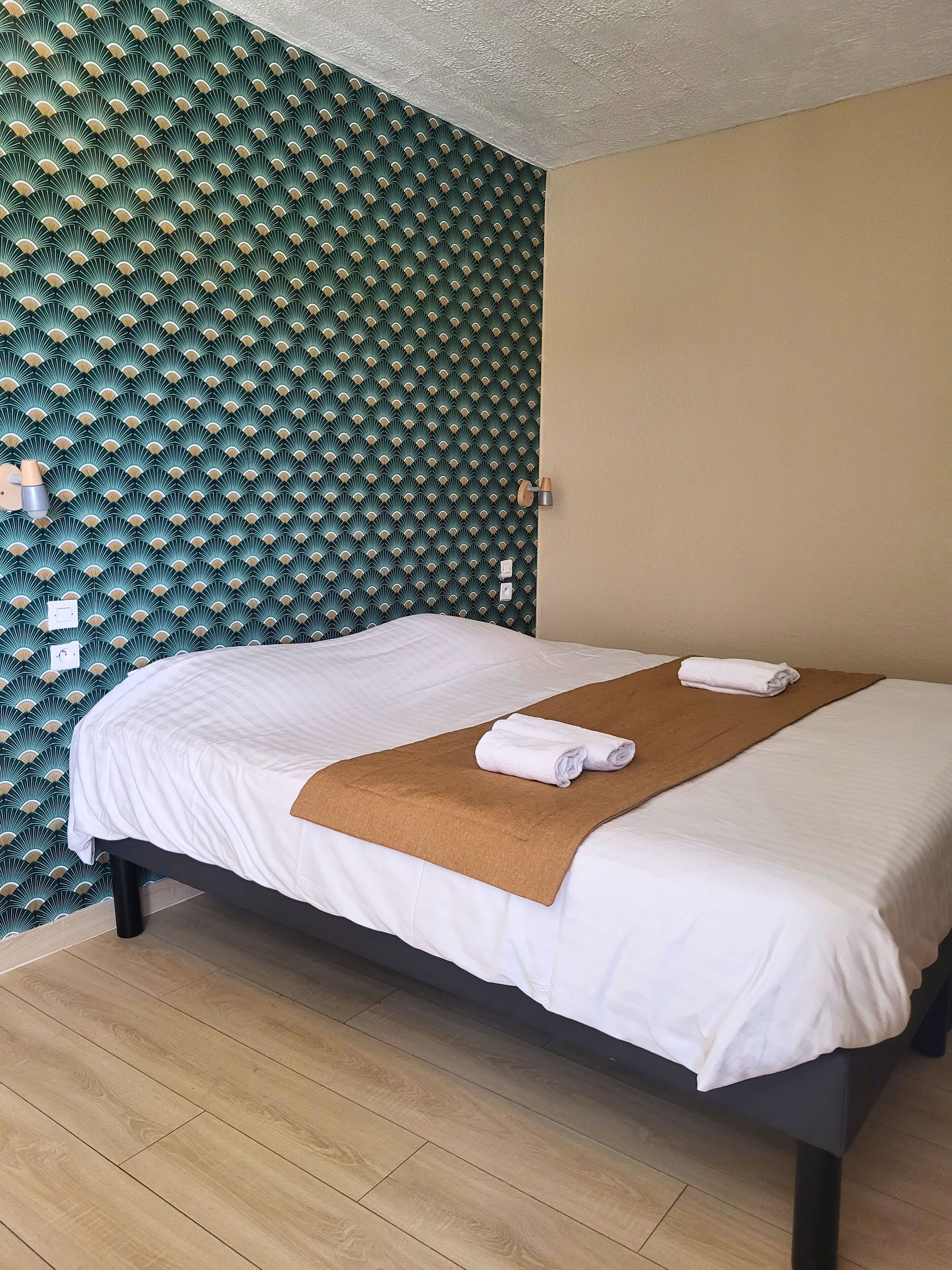 Een keurig opgemaakt bed met witte lakens en een bruine plaid, een houten vloer, en een groene, behangende wand met een gestructureerd plafond.