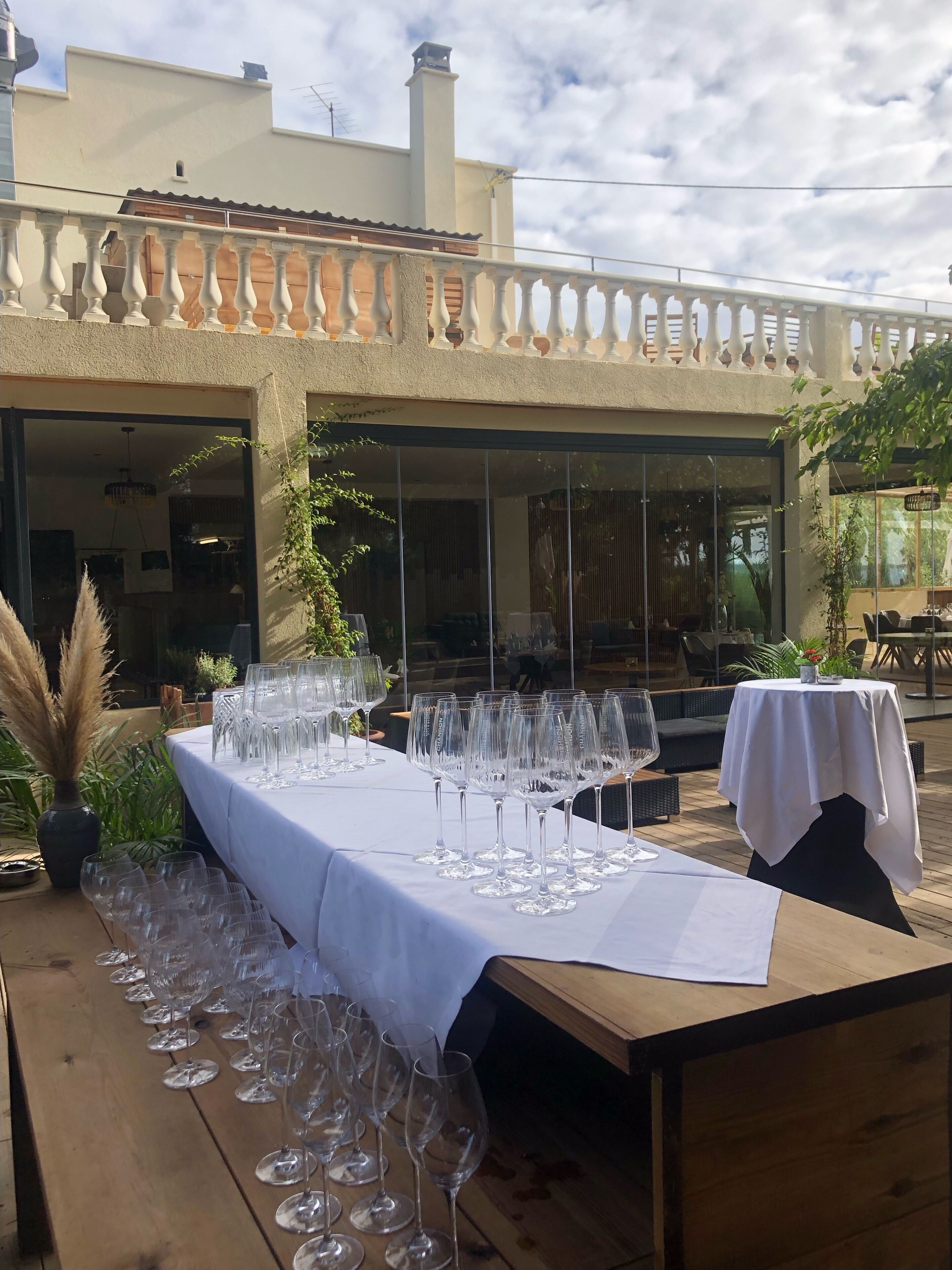 Un área de comedor al aire libre con una larga mesa dispuesta con copas de vino, rodeada de plantas en macetas y un banco de madera.