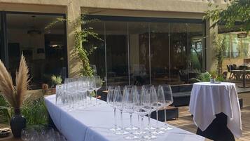 Un área de comedor al aire libre con una larga mesa dispuesta con copas de vino, rodeada de plantas en macetas y un banco de madera.