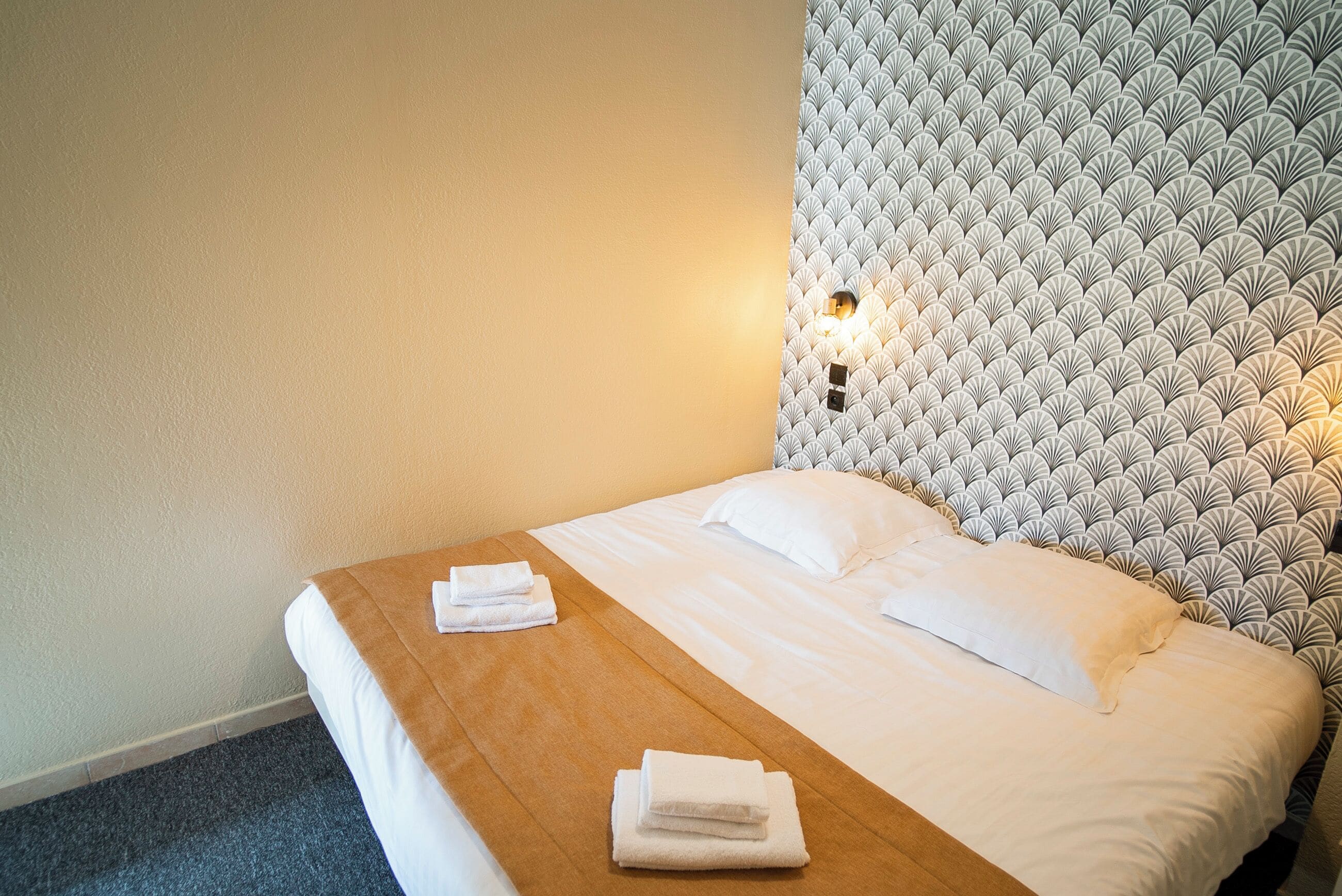 A neatly made bed with white linens and a brown headboard, a wooden bedside table with folded towels, and a patterned wall with a light fixture.