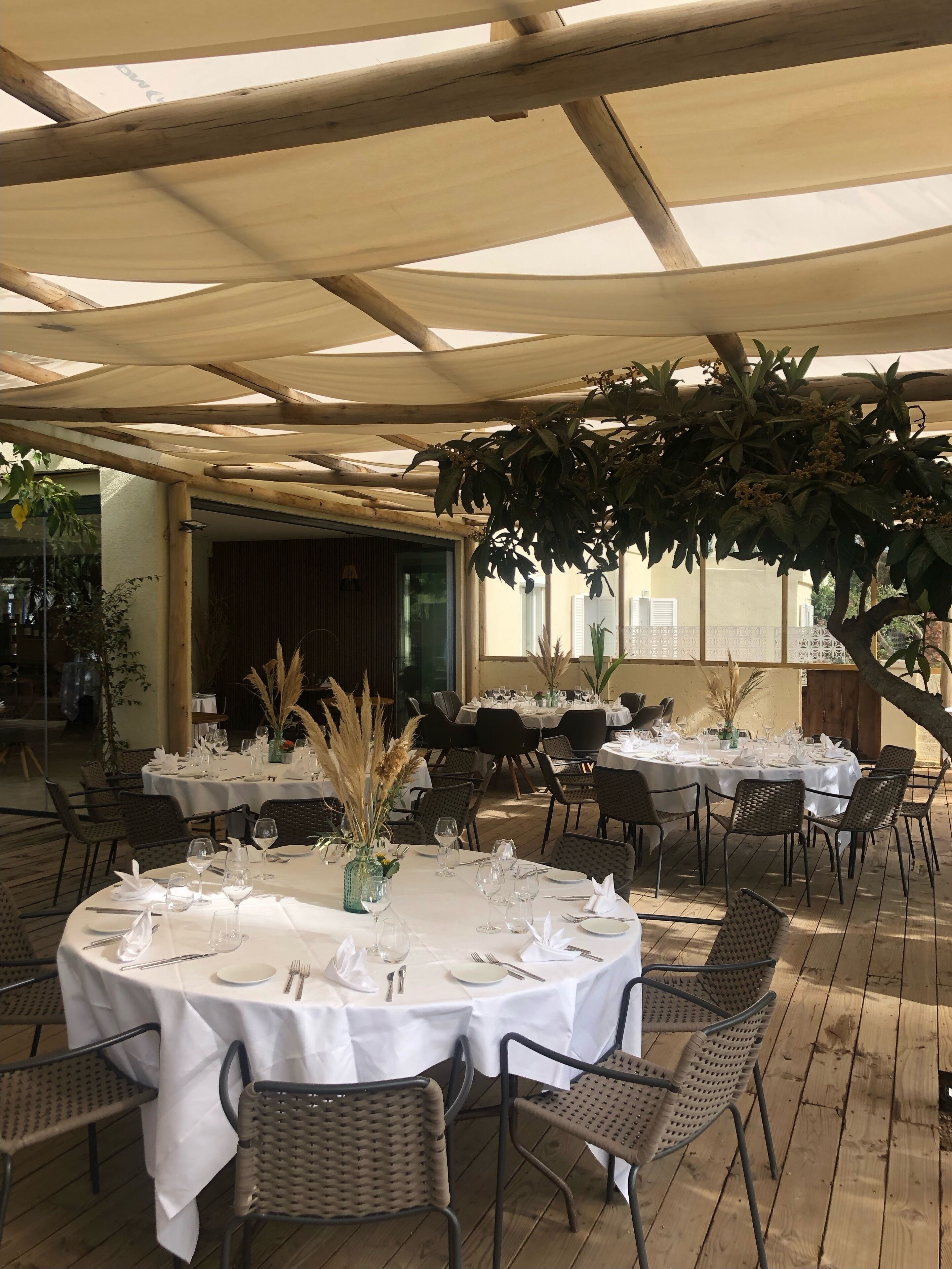 An outdoor dining area with round tables set for two, covered by a wooden pergola with a white canopy.