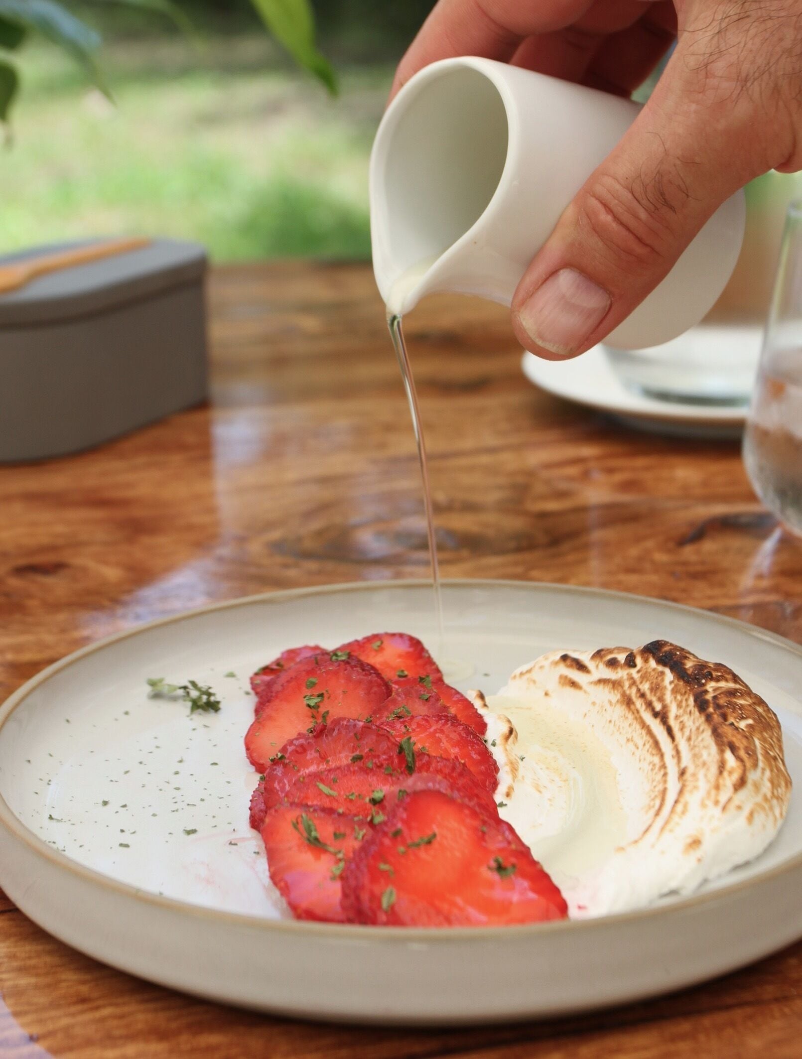 A hand pouring a liquid onto a plate with strawberries and a meringue.