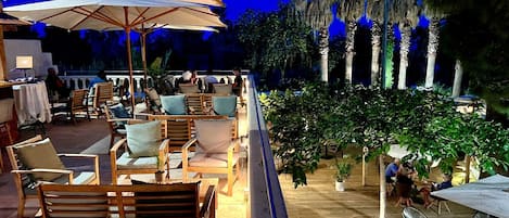 An outdoor seating area with wooden furniture and umbrellas, surrounded by palm trees and potted plants.