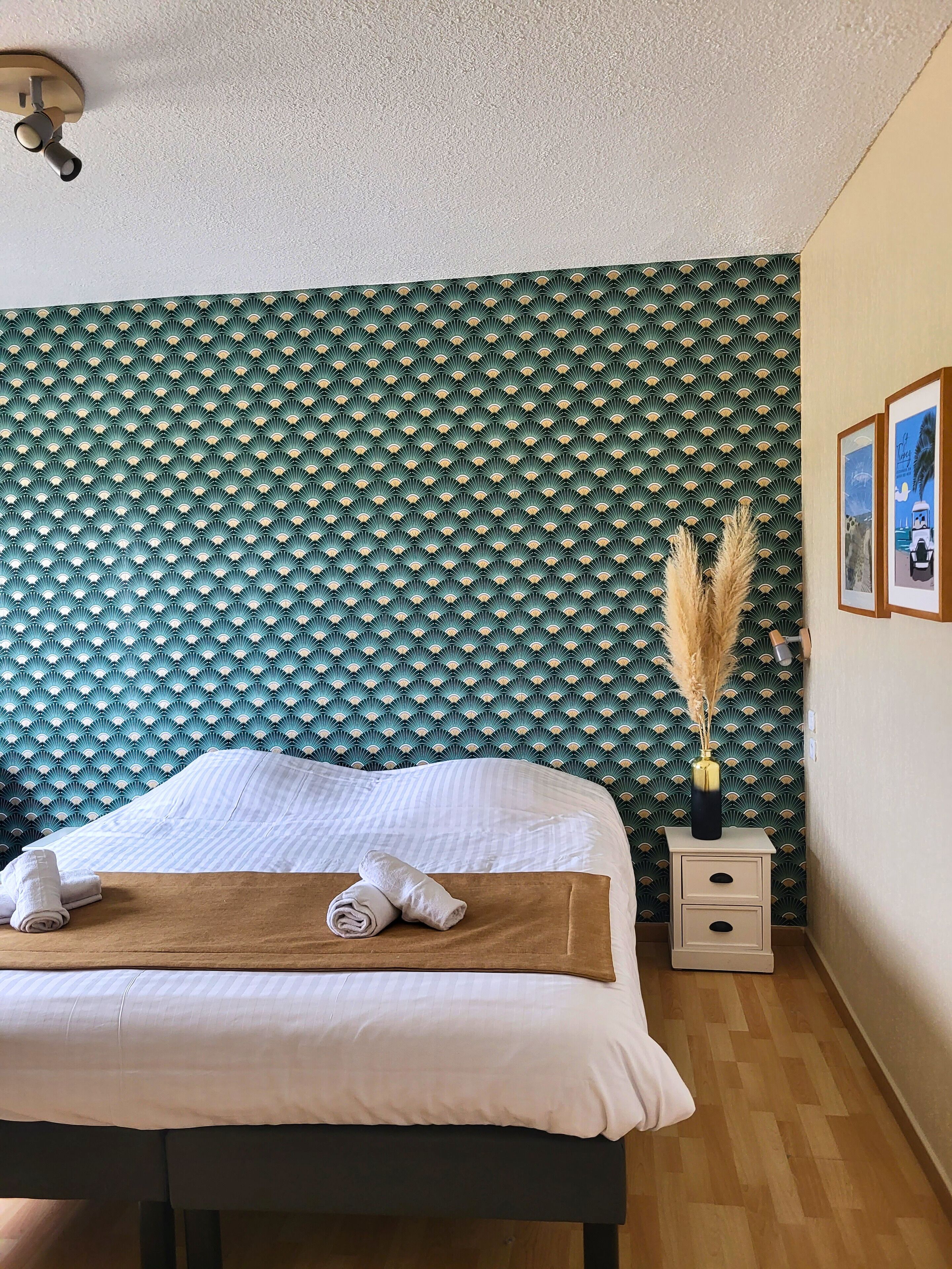 Una cama bien hecha con sábanas blancas y una colcha marrón, una mesita de noche con un florero de plumas de pampa, y una pared con papel tapiz estampado en verde y blanco.