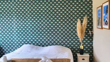 Una cama bien hecha con sábanas blancas y una colcha marrón, una mesita de noche con un florero de plumas de pampa, y una pared con papel tapiz estampado en verde y blanco.