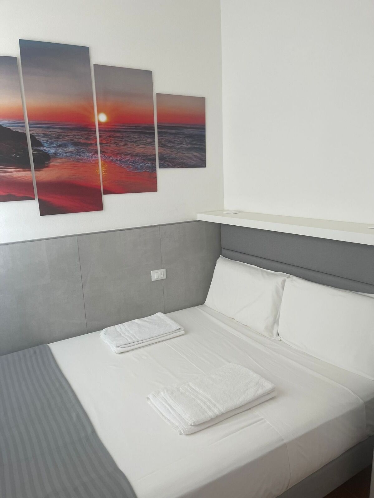 A neatly made bed with white linens and a folded towel, a grey headboard, and three sunset paintings on the wall.