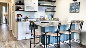 Una cocina moderna con armarios blancos, encimera de granito y taburetes de bar. Hay estantes sobre la encimera con platos y vasos. Un reloj y un cartel que dice "The Beach is calling and I must go" están en la pared.