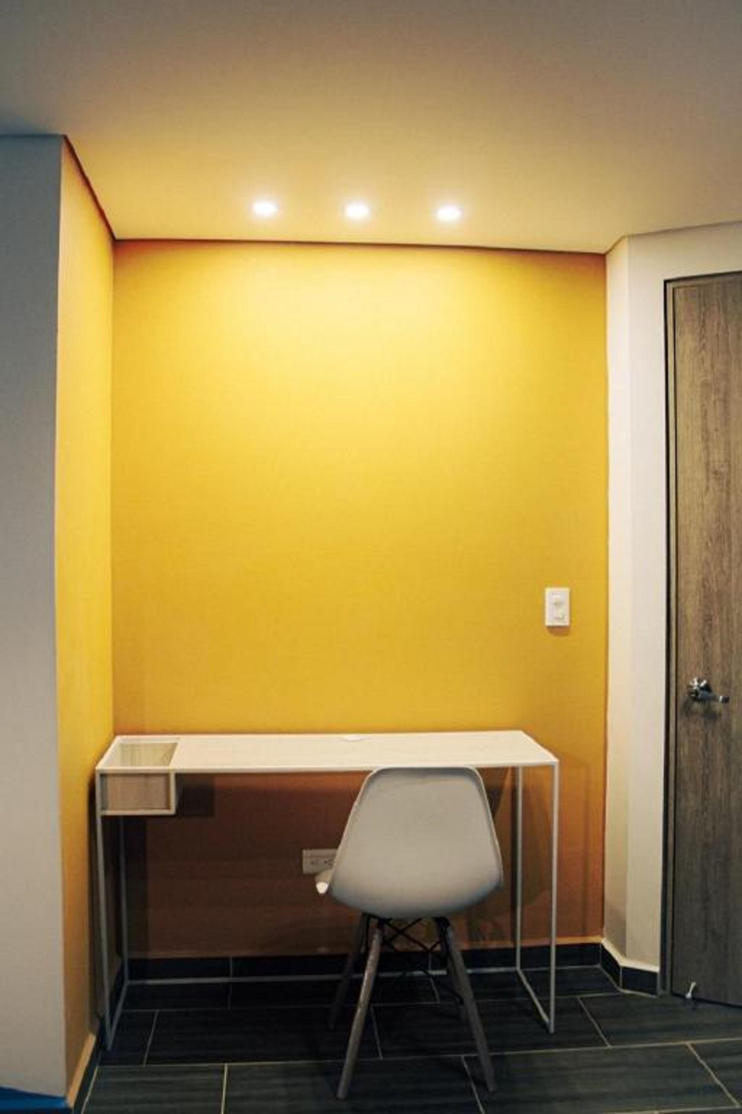 Una habitación pequeña y cerrada con un escritorio y una silla blancos, una puerta de madera y una pared amarilla.