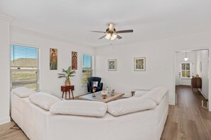 Un salon spacieux avec un grand canapé d’angle blanc, un ventilateur de plafond et des œuvres d’art accrochées aux murs.