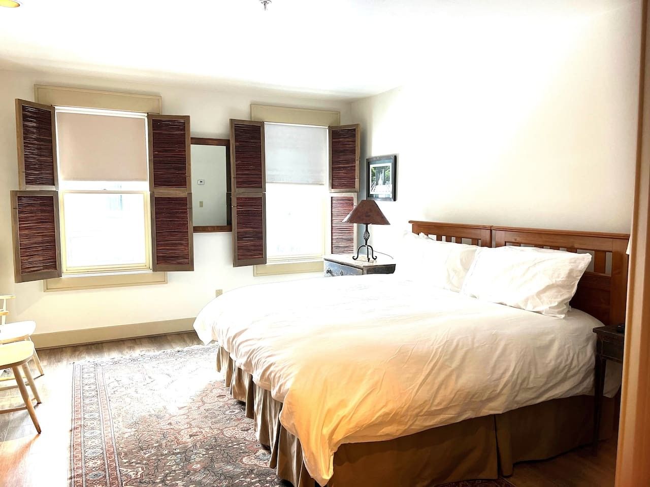 A bedroom with a large bed, a desk, and a chair. There are two windows with shutters, a lamp, and a framed picture on the wall.