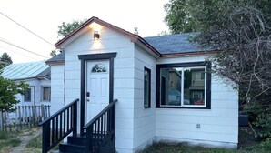 A small, single-story house with a white exterior and black trim. It features a front door with a small window above it, a staircase with black railings leading to the entrance, and a small porch area.