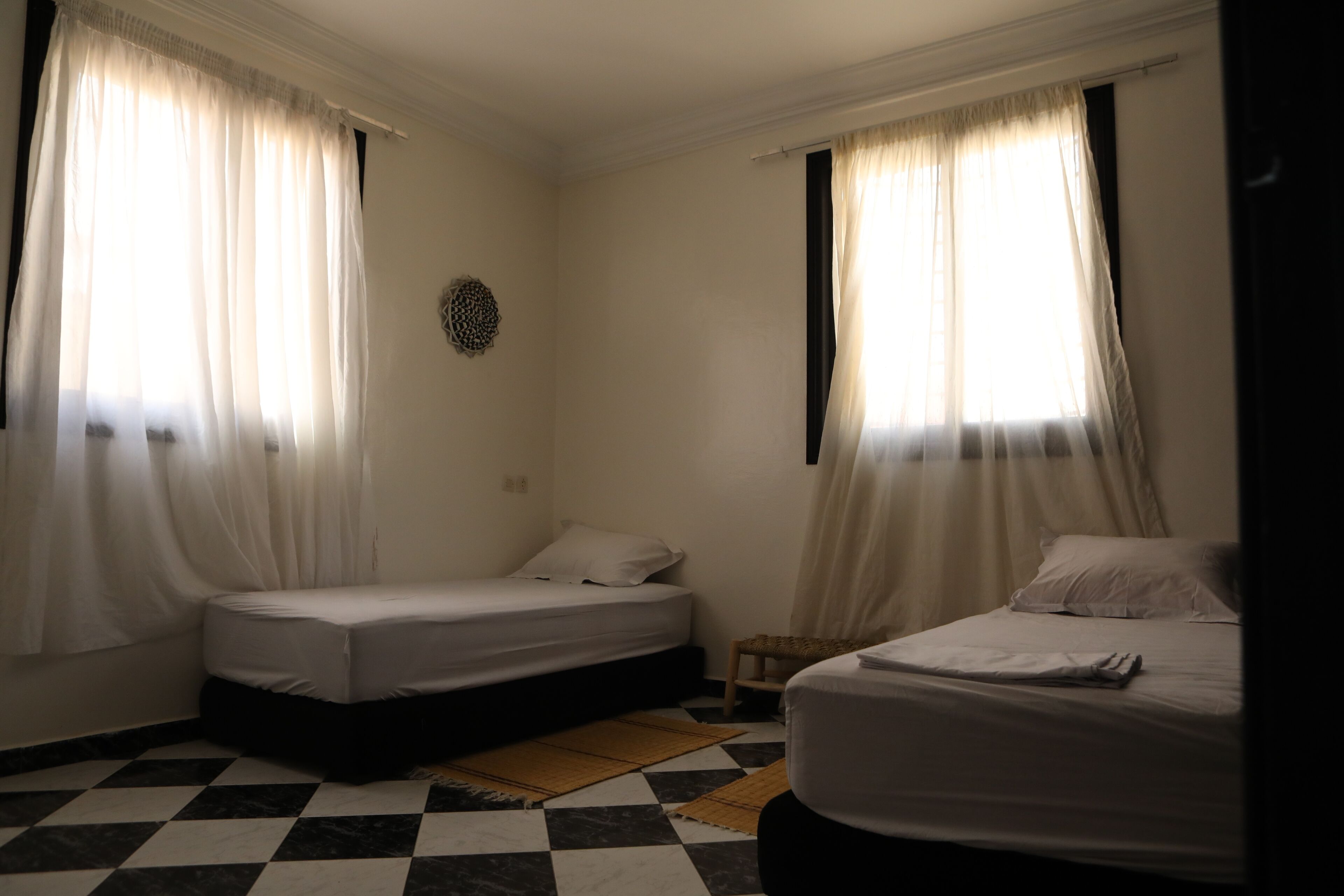 Een kamer met twee bedden, een geruit vloertje en dunne gordijnen.