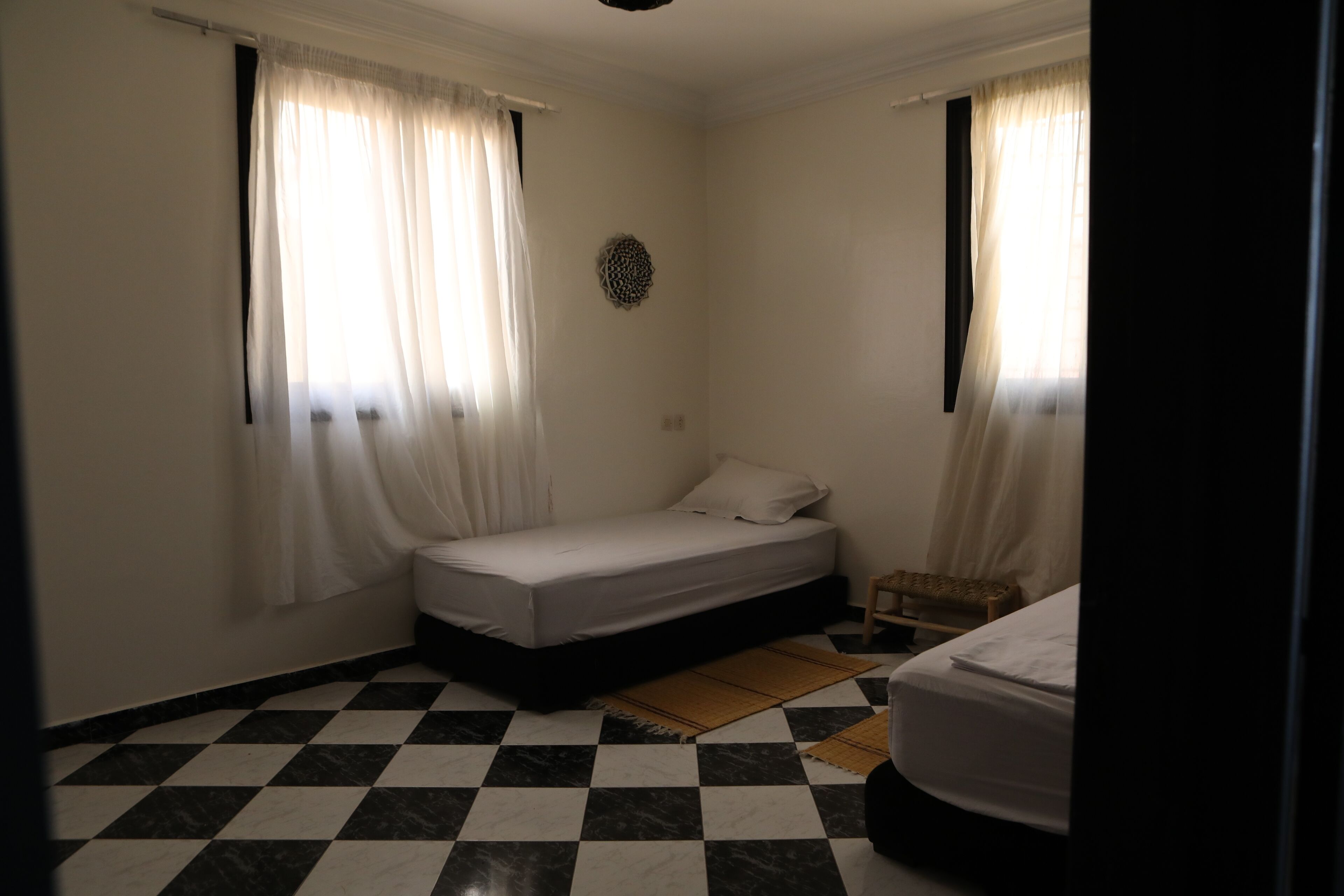 Ein kleiner, sauberer Raum mit einem Schachbrettmuster auf dem Boden, einem Einzelbett, einem kleinen Tisch und zwei Fenstern mit durchsichtigen Vorhängen.