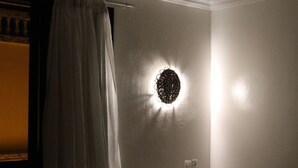 Une petite pièce faiblement éclairée, avec un lit simple, un luminaire fixé au mur et une fenêtre avec des rideaux.