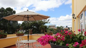 Een terras met een ronde tafel en stoelen, een beige parasol en plantenbakken met roze bloemen.