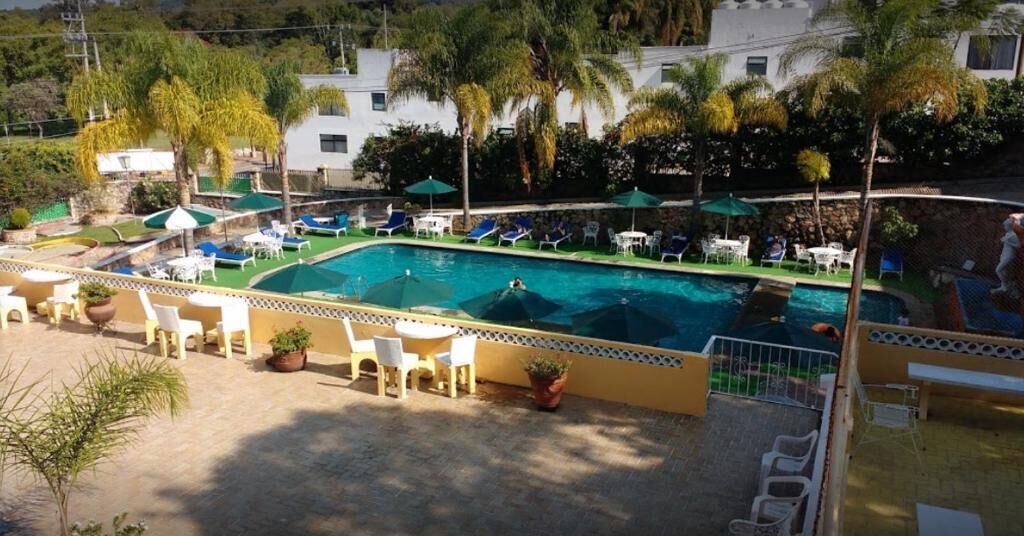 A pool area with lounge chairs and umbrellas, surrounded by palm trees and a building in the background.