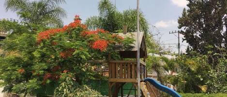 A wooden play structure with a slide, surrounded by lush greenery and red flowers.
