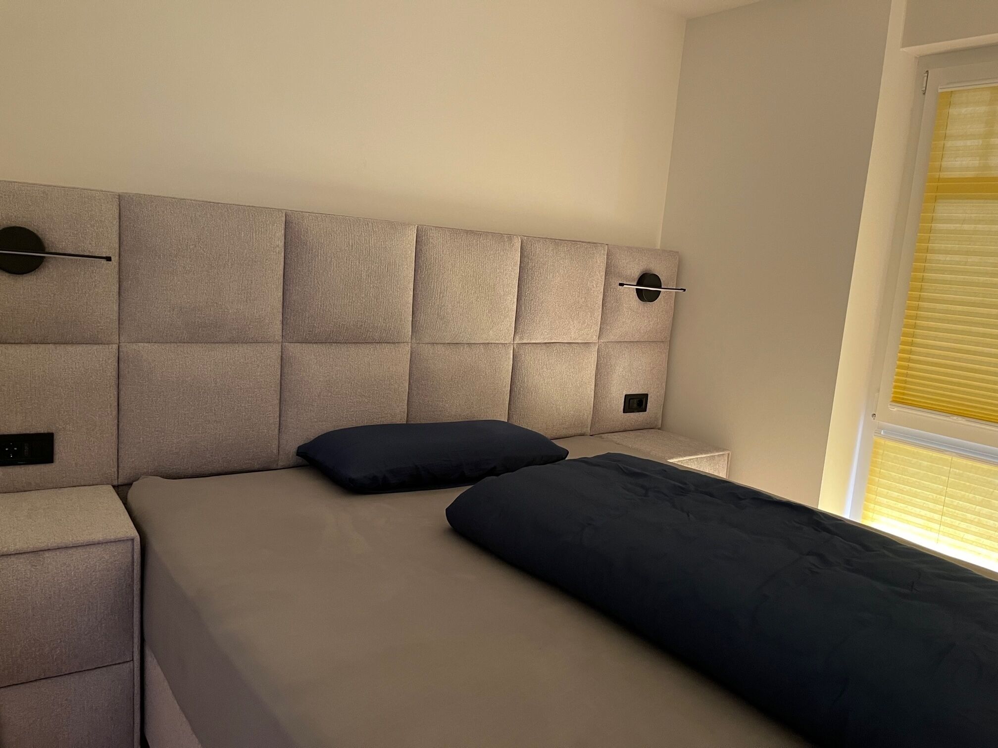A modern bedroom with a tufted headboard, a bed with dark bedding, a bedside table, and a window with blinds.