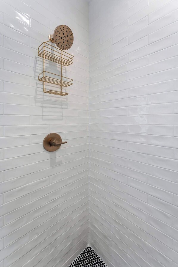 Esquina de un baño con azulejos blancos tipo metro, un cabezal de ducha dorado y un perchero dorado.