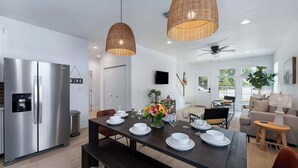 A modern kitchen with stainless steel appliances, a dining area with a dark wooden table set for four, and a living area with a sofa and a lamp. - Luxury Escape at The Noir: 3br, Hot Tub, & Steps From Main St! (Fredericksburg)