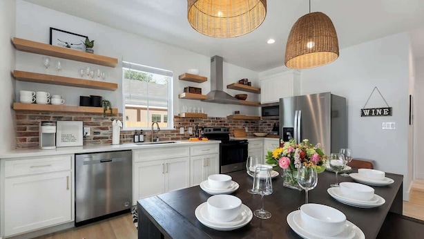 A modern kitchen with white cabinets, a stainless steel dishwasher, and a dining table set with white plates and glasses. - Luxury Escape at The Noir: 3br, Hot Tub, & Steps From Main St! (Fredericksburg)