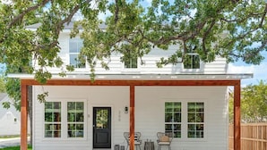 A white house with a covered porch, two windows, a door, and a small seating area with chairs and a table. - Luxury Escape at The Noir: 3br, Hot Tub, & Steps From Main St! (Fredericksburg)