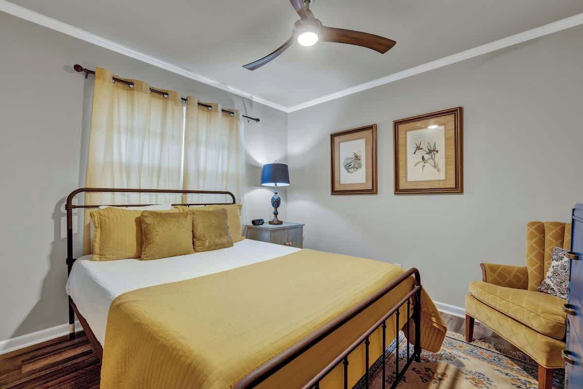 A bedroom with a bed, a ceiling fan, a nightstand, a lamp, a chair, and two framed pictures on the wall.