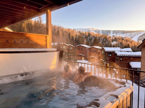 Un jacuzzi con dos personas, cabañas de madera al fondo y un paisaje nevado de montaña.
