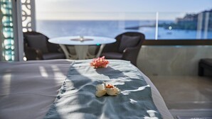 Un balcone con tavolo e sedie, un letto con un vaso di fiori e vista sul mare e sugli edifici.