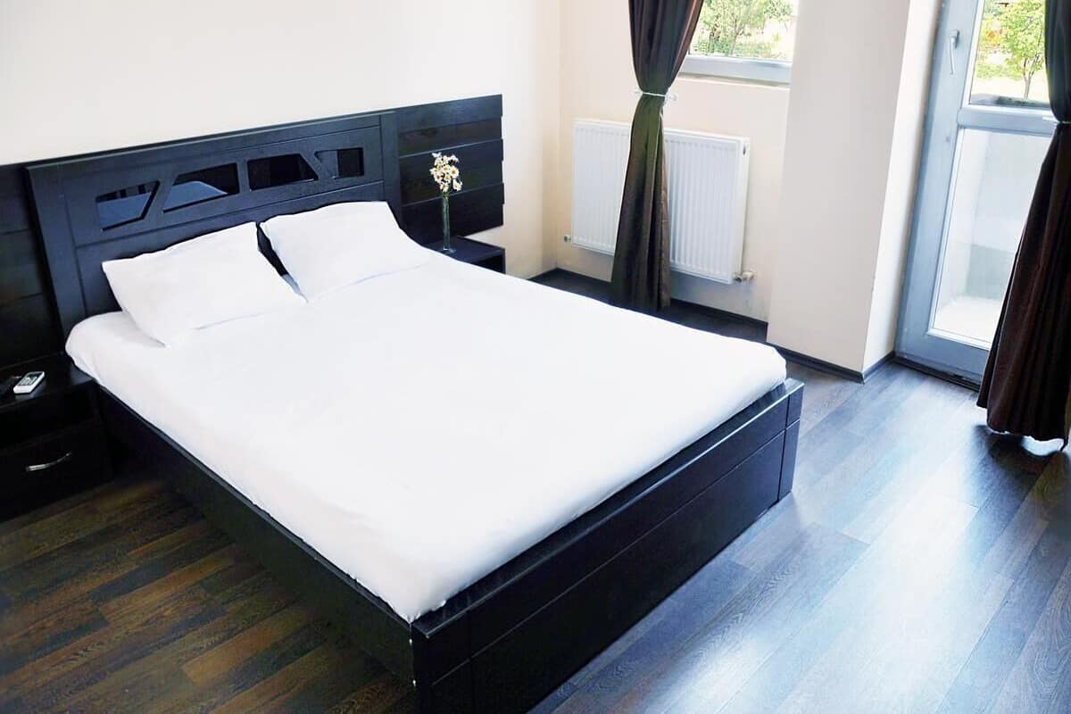 A modern bedroom with a large bed, dark wood flooring, a nightstand, a bedside table, and a window with curtains.