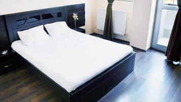 A modern bedroom with a large bed, dark wood flooring, a nightstand, a bedside table, and a window with curtains.