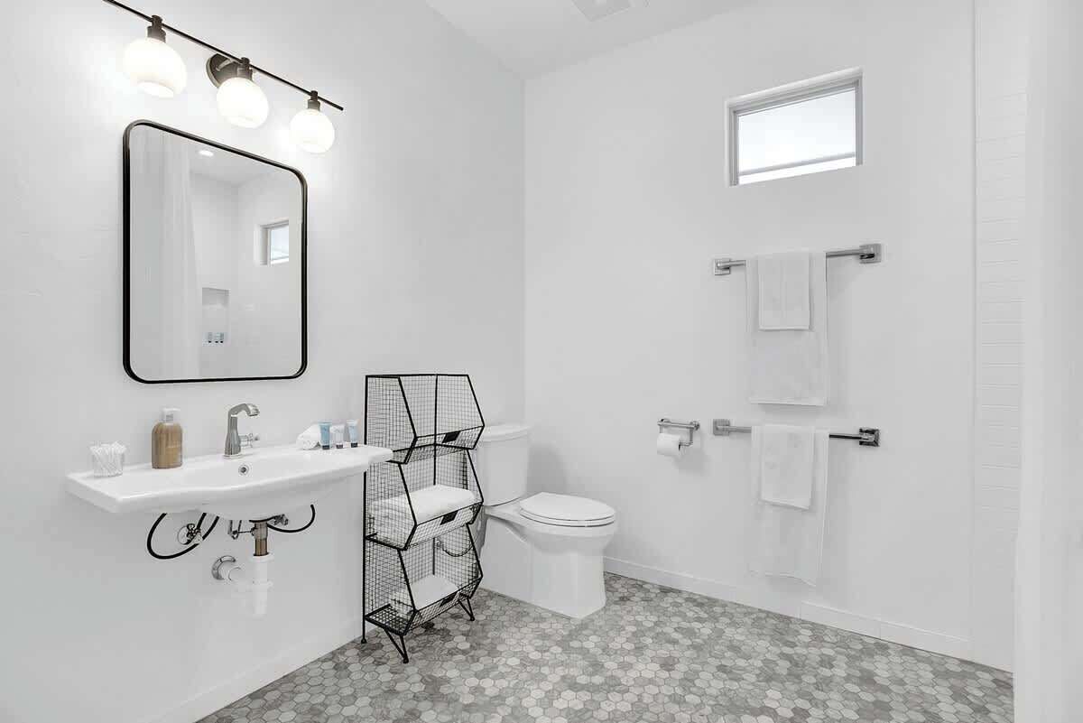 A modern bathroom with a white sink, mirror, and toilet. There is a window above the toilet and a towel rack on the wall.