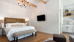 Une chambre avec une tête de lit en bois, un lit avec une literie blanche, un banc, un lustre, un téléviseur fixé au mur et une petite cuisine avec une étagère.