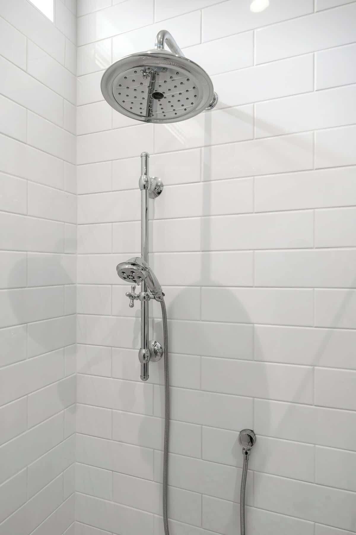 A shower with a rain showerhead and a handheld showerhead, mounted on a wall with white tiles.