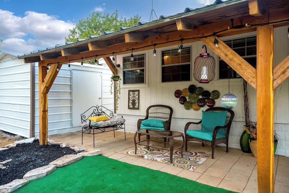 A covered patio area with wooden beams, two wicker chairs with teal cushions, a patterned rug, and decorative hanging lights.