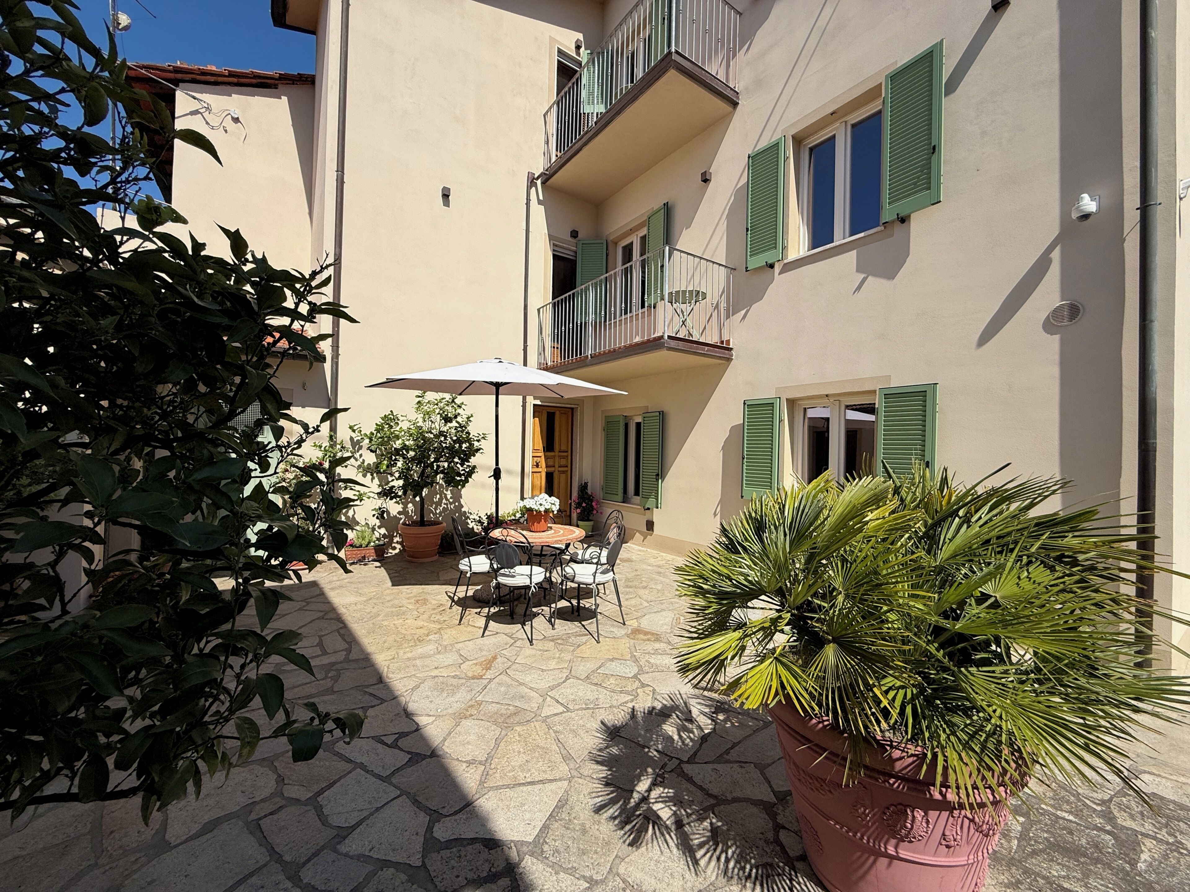 Un patio con una mesa y sillas, plantas en macetas y un edificio con balcones y contraventanas.