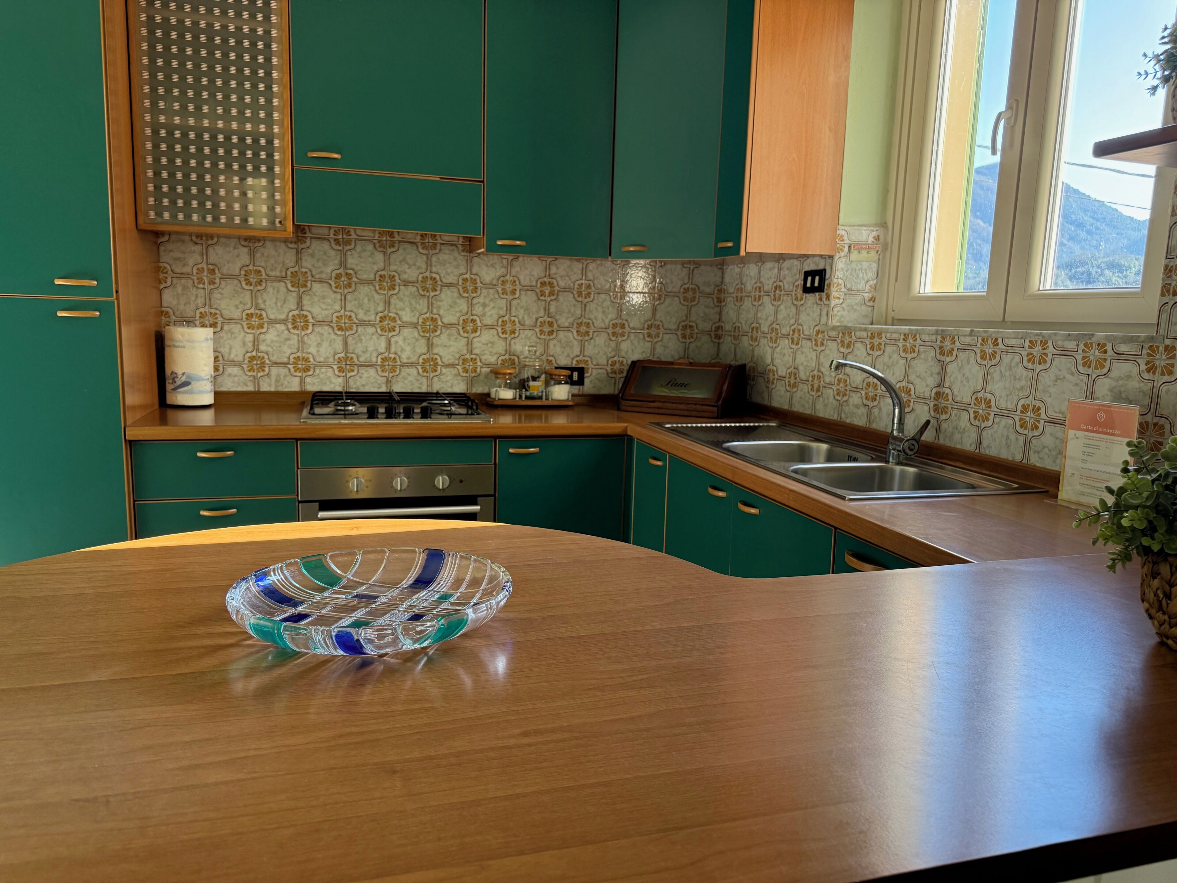 A kitchen with teal cabinets, a wooden countertop, a glass bowl, a stove, and a window with a view.
