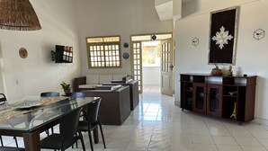 A dining area with a glass table, black chairs, a brown sofa, and a wicker chair. There is a TV mounted on the wall and a cabinet with decorative items.