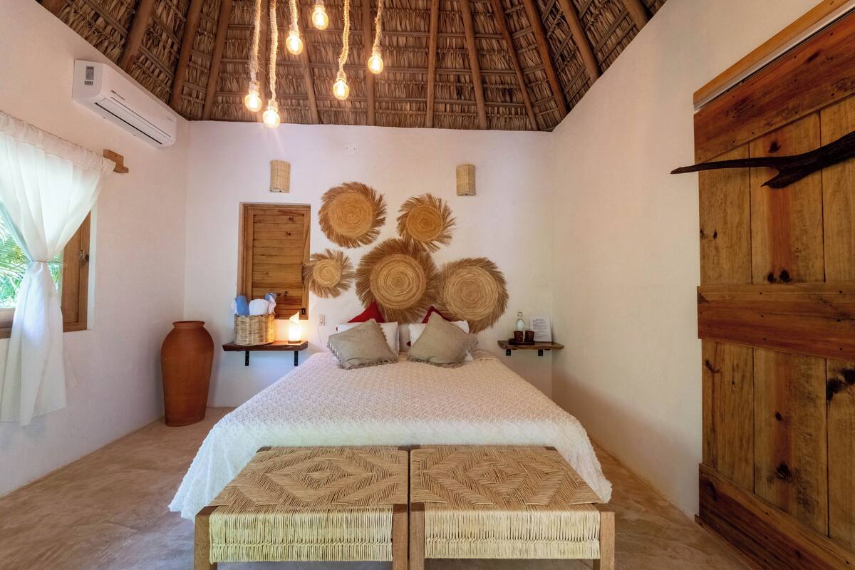 A bedroom with a thatched roof, a bed with a white cover, woven headboards, and a rustic wooden wardrobe.