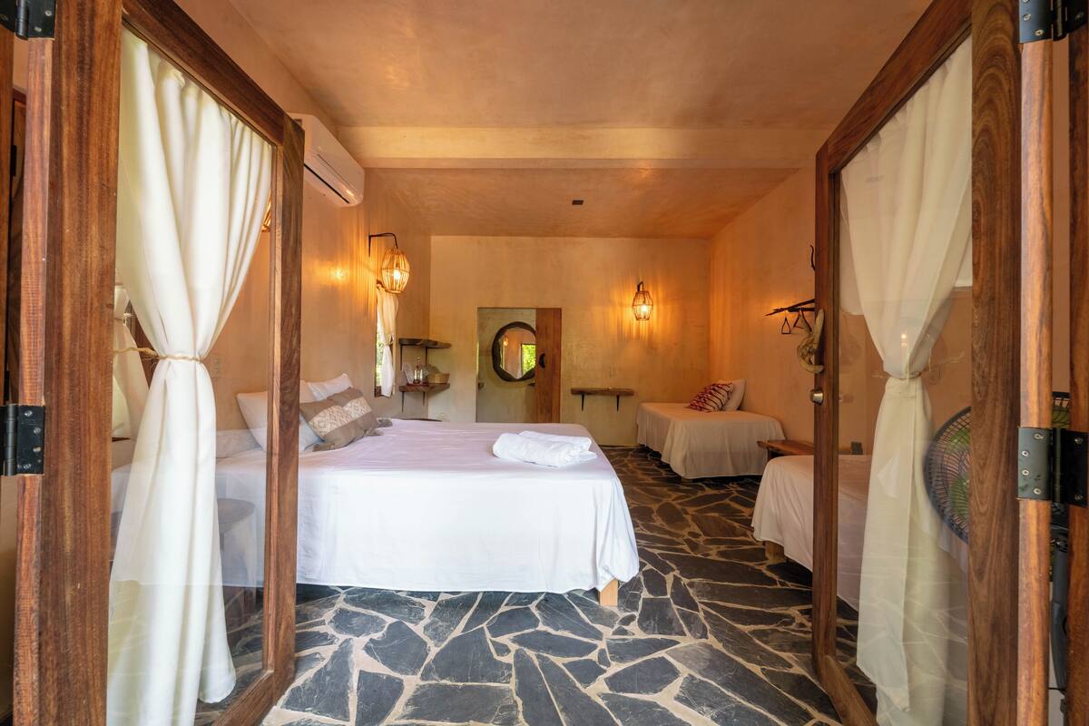 A hotel room with a large bed, two smaller beds, stone flooring, wooden accents, and a mirror with curtains.
